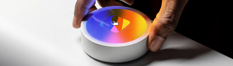 Main sur un bouton circulaire orné du symbole A Kind of Magic et d’un dégradé multicolore.