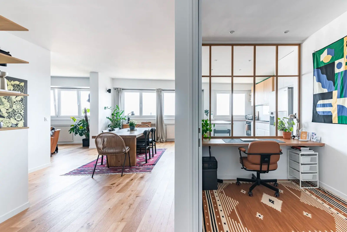 Appartement parisien lumineux avec parquet clair, salle à manger contemporaine et bureau séparé par une verrière en bois, espace de travail minimaliste et plantes vertes.