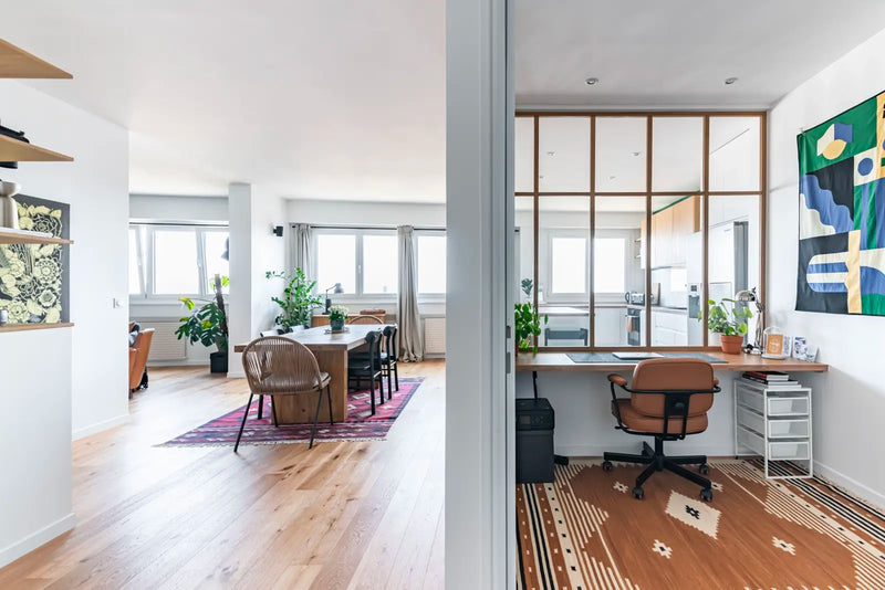 Appartement parisien lumineux avec parquet clair, salle à manger contemporaine et bureau séparé par une verrière en bois, espace de travail minimaliste et plantes vertes.