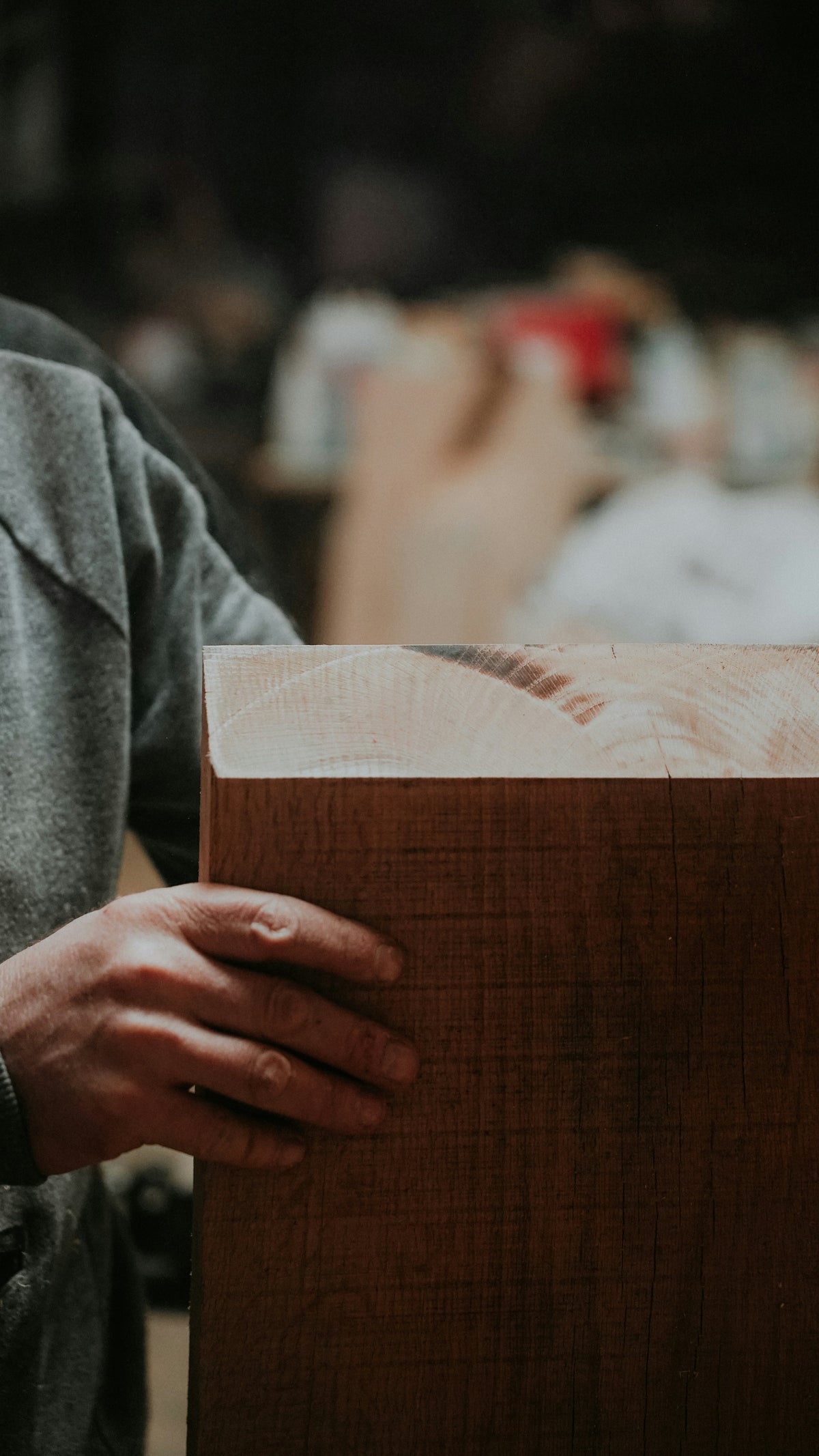 Main d'un artisan tenant une pièce de bois massif aux veines apparentes, dans un atelier de menuiserie avec l'arrière-plan flou montrant l'activité de travail du bois.