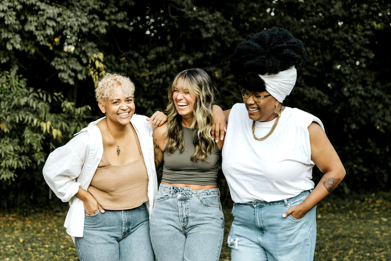 Trois amies souriantes debout dans un jardin, les bras autour des épaules.