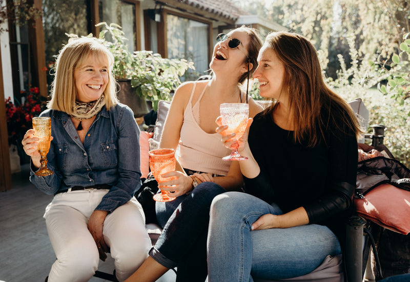 Trois amies assises en terrasse rient ensemble, un verre à la main.