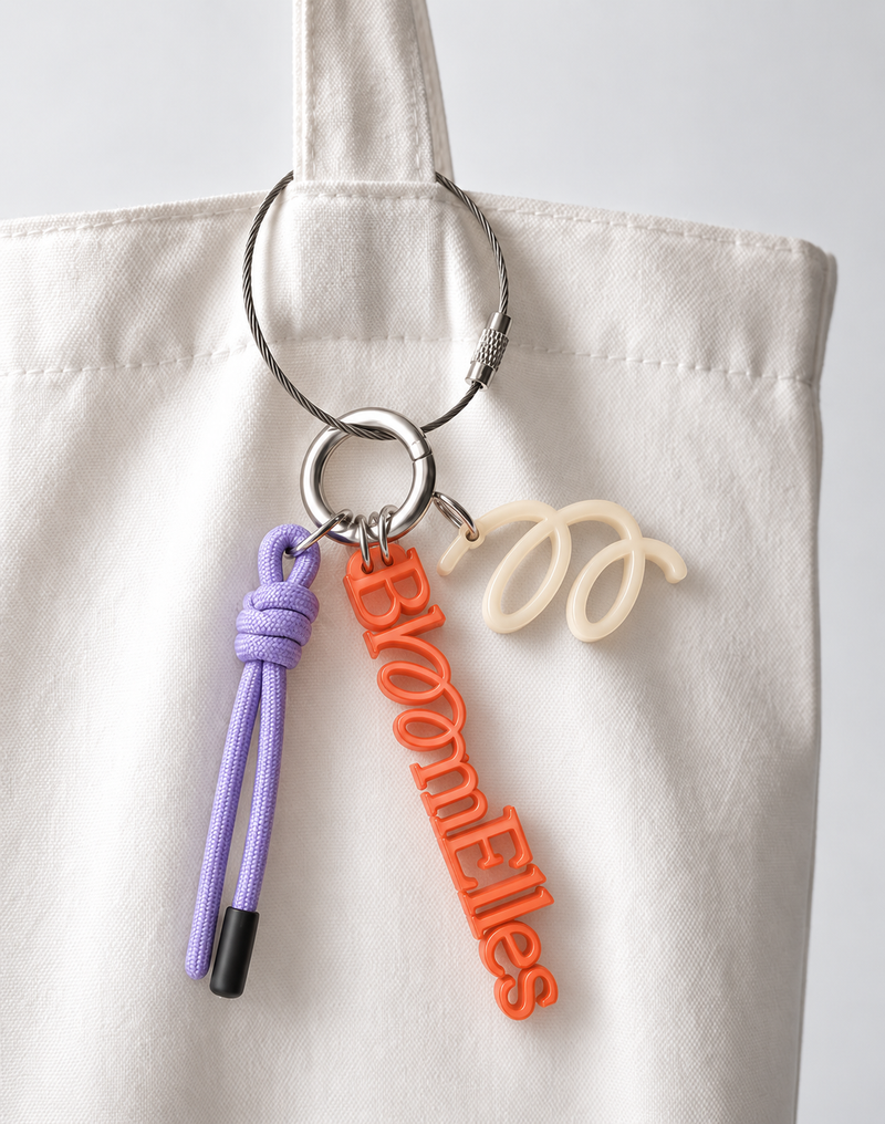 Porte-clés BloomElles orange, avec cordon lavande et breloque crème, accroché à un sac blanc.
