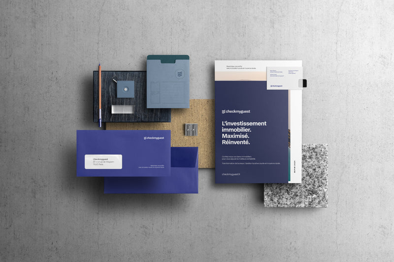 Flatlay de l'identité visuelle Checkmyguest : enveloppes bleu marine, carte de visite, affiche 'L'investissement immobilier maximisé' et échantillons de matériaux.
