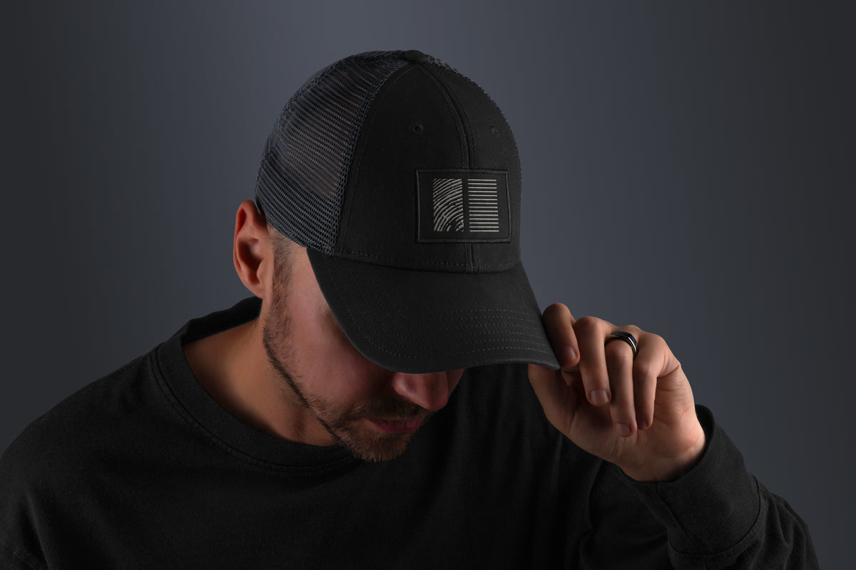 Portrait d’un homme portant une casquette noire avec un logo rectangulaire à lignes graphiques, tête légèrement baissée, fond gris uni et lumière douce, style moderne et discret.