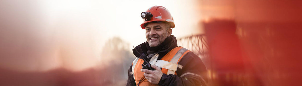Ouvrier sur un chantier de construction portant un casque de sécurité et un gilet haute visibilité, utilisant une radio sur un site industriel.