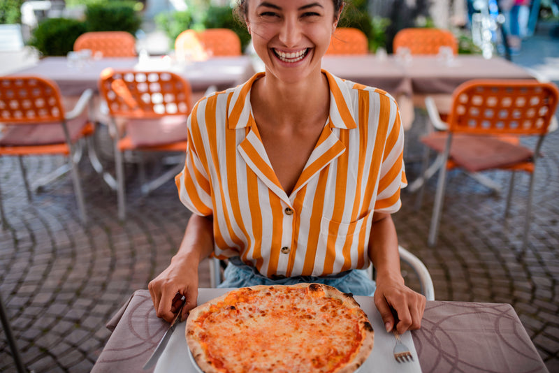 Femme souriante assise en terrasse de restaurant devant une pizza prête à être dégustée.