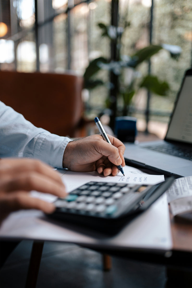 Gros plan sur des mains prenant des notes manuscrites à côté d'une calculatrice et d'un ordinateur portable, dans un bureau lumineux.