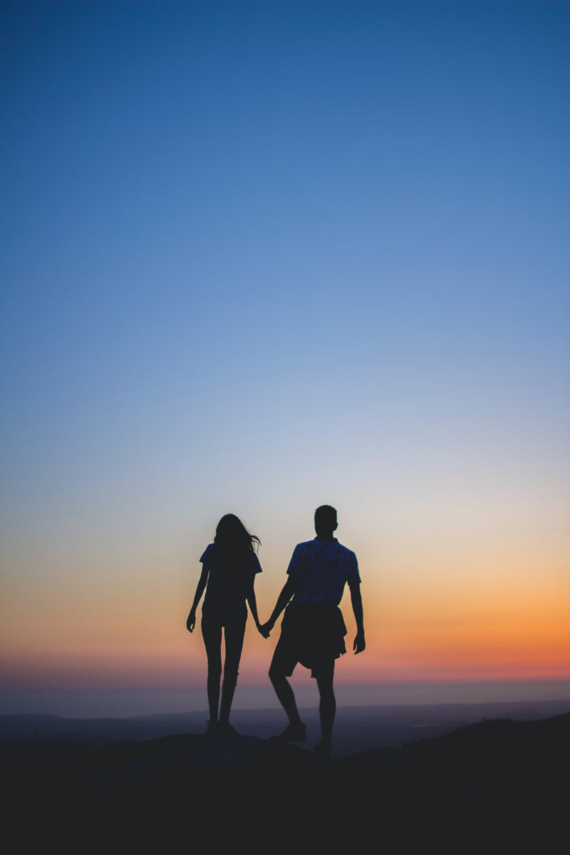Silhouette d’un couple se tenant la main au sommet d’une colline au coucher du soleil, symbole de relation et de projet de vie.