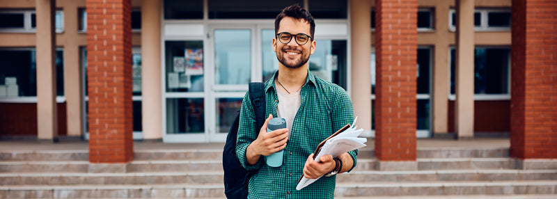 Étudiant souriant devant un campus universitaire avec sac et documents, illustrant les études en management et commerce de la santé.