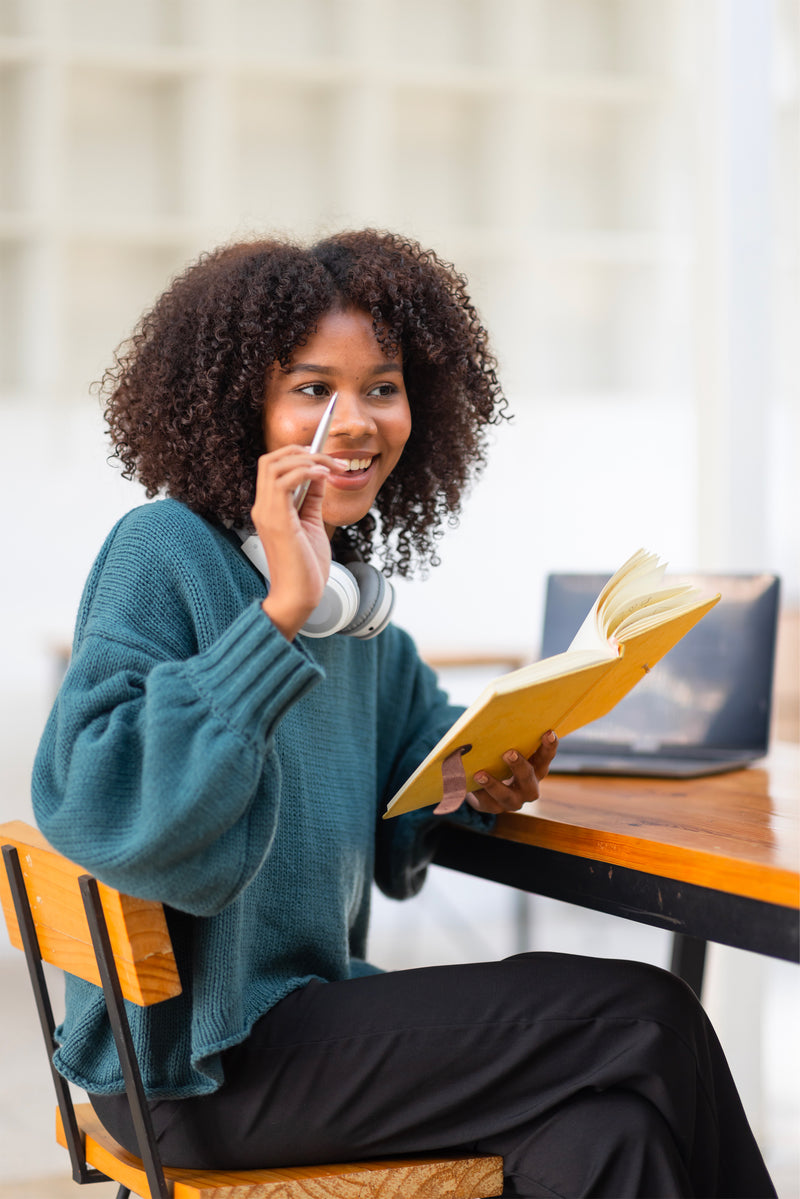 Étudiante souriante prenant des notes dans un carnet pendant ses études, avec casque audio autour du cou dans un espace de travail.