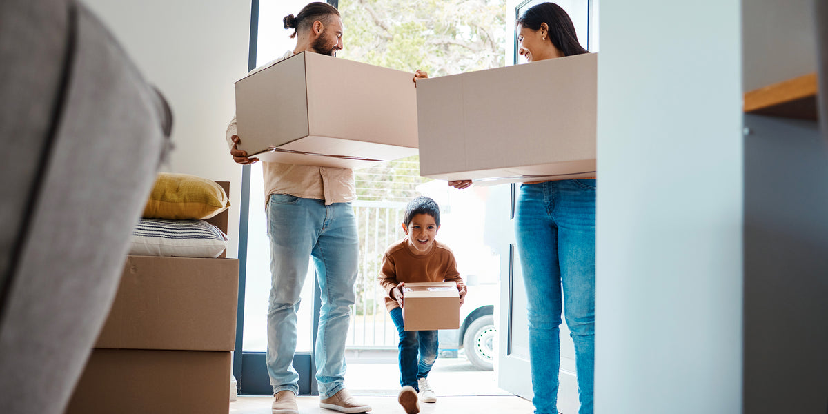 Famille avec enfant entrant dans une nouvelle maison en portant des cartons lors d’un déménagement après un achat immobilier.