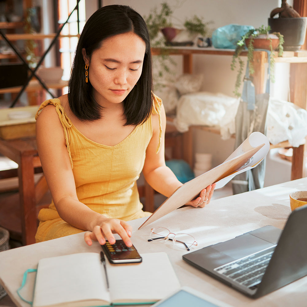 Femme utilisant un smartphone comme calculatrice pour gérer ses finances personnelles et consulter des documents à son bureau.