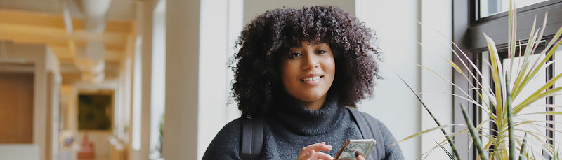 Portrait d’une femme souriante aux cheveux bouclés, près d’une fenêtre et d’une plante.