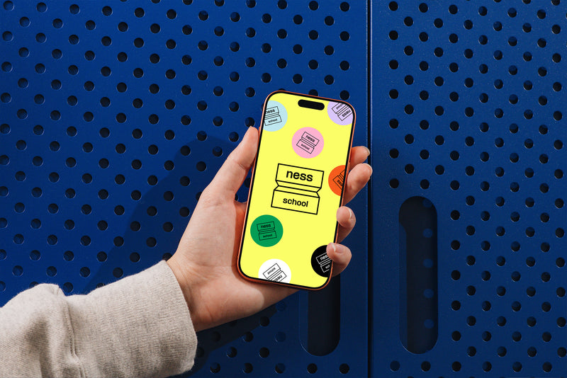 Smartphone tenu en main affichant le logo Ness School et ses déclinaisons colorées sur fond jaune.