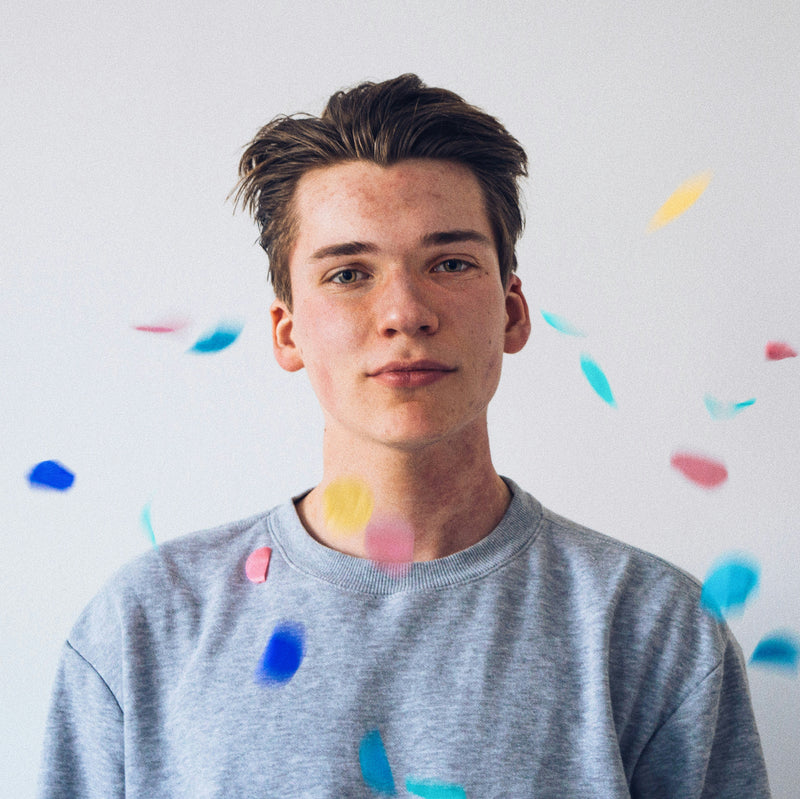 Portrait d’un jeune homme en tee-shirt gris, entouré de confettis colorés.