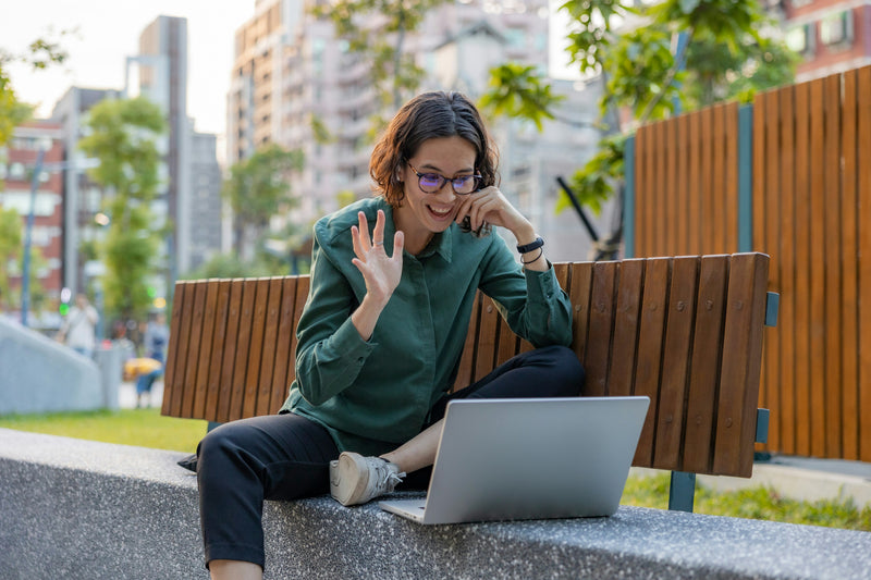 Femme assise en extérieur devant un ordinateur portable, saluant de la main pendant un appel vidéo.