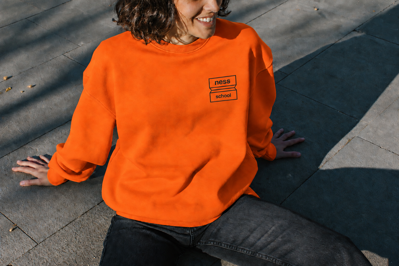 Sweat orange avec le logo Ness School imprimé en noir sur la poitrine, porté par une personne assise.