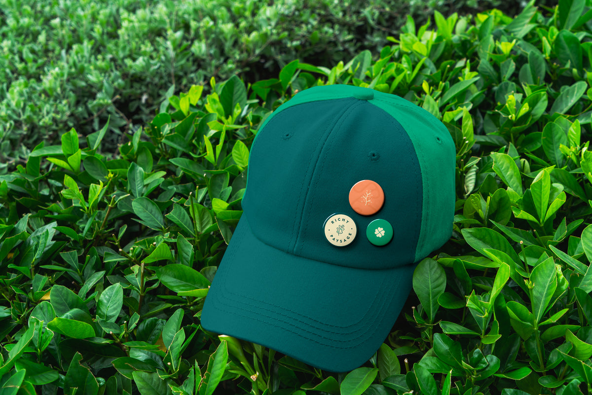 Casquette verte avec badges Richy Paysage posée sur un feuillage dense, illustrant l’identité visuelle d’un paysagiste et son univers naturel.