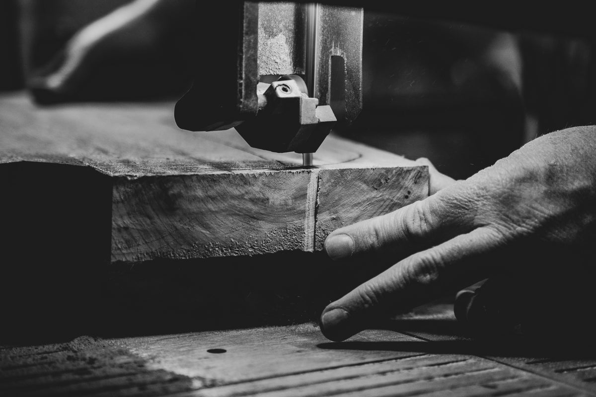 Photo en noir et blanc d’un artisan qui guide une planche de bois sous une scie à ruban, mettant en valeur le geste précis de la main et la texture du bois.