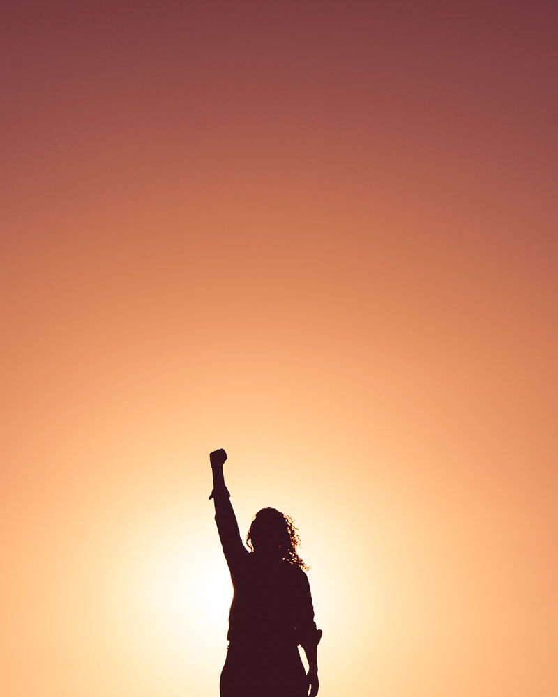 Silhouette d’une femme levant le poing au coucher du soleil, symbole de force, espoir et empowerment.
