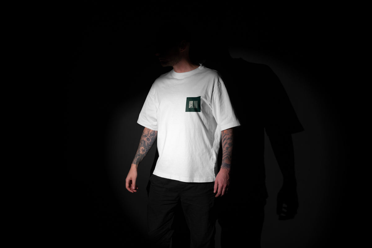 Homme de profil portant un t-shirt blanc oversize avec un petit carré vert imprimé sur la poitrine gauche, fond noir dramatique, tatouages visibles sur les deux avant-bras.