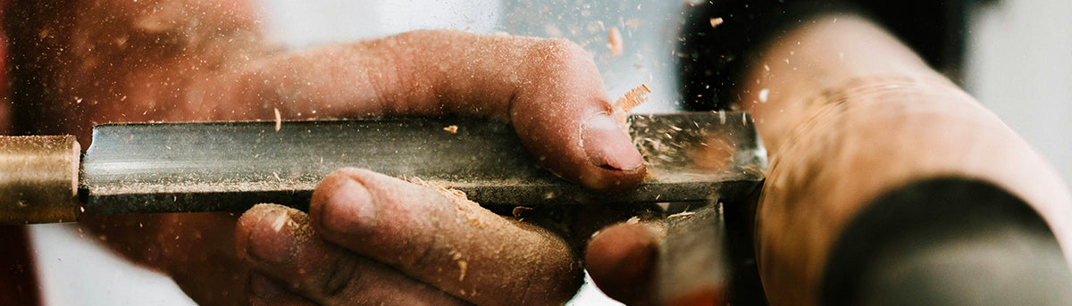 Mains d'un artisan tenant un burin contre une pièce de bois en rotation sur un tour, avec des copeaux et de la sciure projetés en l'air