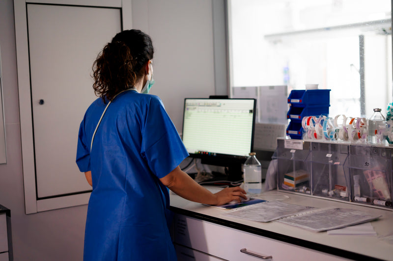 Soignante masquée de dos travaillant sur un poste informatique en service hospitalier.